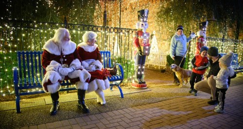 Two Santa figures sit on a bench surrounded by festive lights, while children and a dog interact nearby.
