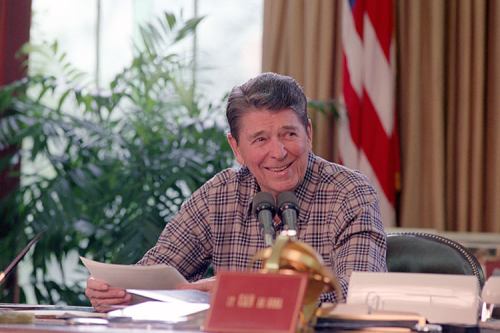 Ronald Reagan smiles while seated at a desk, holding papers, with a U.S. flag and plants in the background.