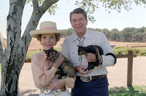 Ronald and Nancy Reagan pose outdoors, each holding a cat, with a tree and open field in the background.