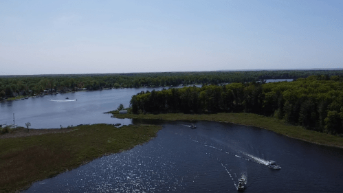 Aerial view of a serene lake surrounded by greenery, with boats gently cruising on the water under a clear blue sky.