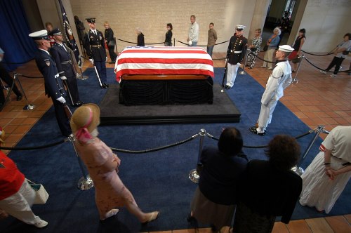A flag-draped casket is surrounded by honor guards and mourners in a solemn setting.