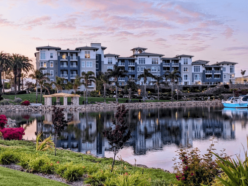 A serene waterfront view of modern apartments with palm trees and colorful flowers, reflecting in the calm water at sunset.