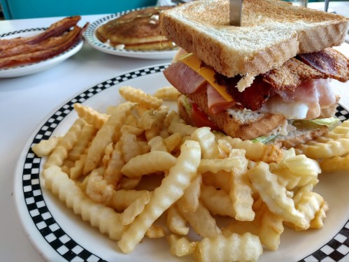 A hearty sandwich stacked with bacon, ham, cheese, and lettuce, served with crinkle-cut fries and pancakes in the background.