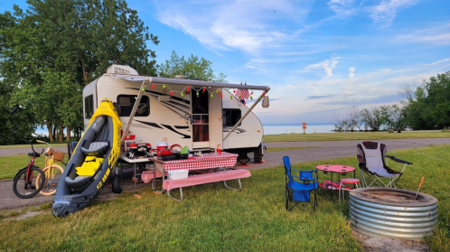 A camper parked by a lake, with outdoor seating, a picnic table, and a kayak nearby, surrounded by greenery.