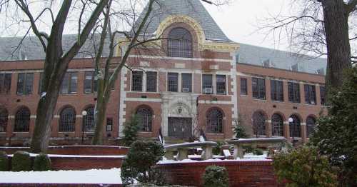 Historic brick building with large windows, surrounded by snow-covered trees and a landscaped courtyard.