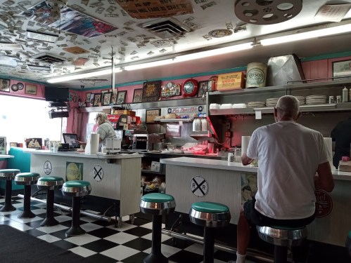 A retro diner interior with checkered floors, vintage decor, and two patrons sitting at the counter.