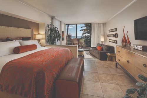 A bright hotel room featuring a king bed, seating area, and large windows with a view of the outdoors.