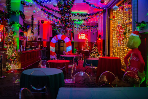 A festive room decorated with colorful lights, Christmas trees, and holiday ornaments, featuring a whimsical Grinch figure.