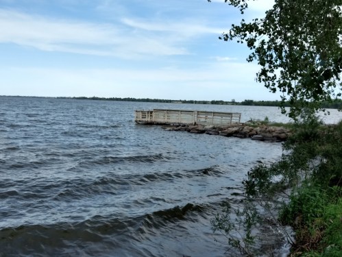A wooden dock extends into a calm lake, surrounded by trees and a clear blue sky. Gentle waves ripple on the water.
