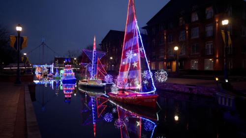 Colorful illuminated boats with festive lights reflect in a calm canal at night, surrounded by buildings and street lamps.