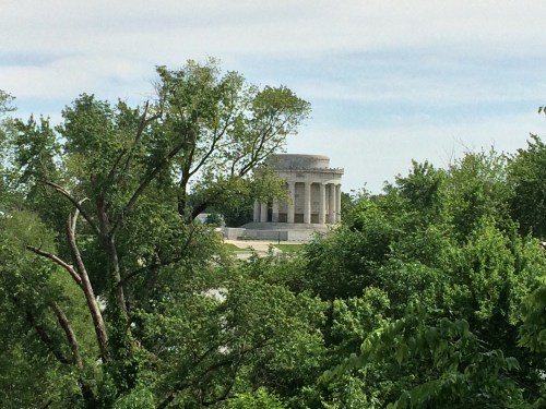 A classical stone monument surrounded by lush green trees under a cloudy sky.