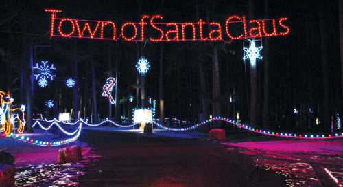 Colorful holiday lights illuminate a dark road, with a sign reading "Town of Santa Claus" and festive decorations.