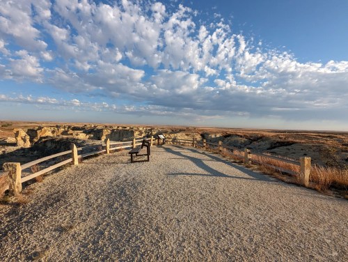 A scenic viewpoint with a gravel path, wooden bench, and a vast sky filled with clouds over a rugged landscape.