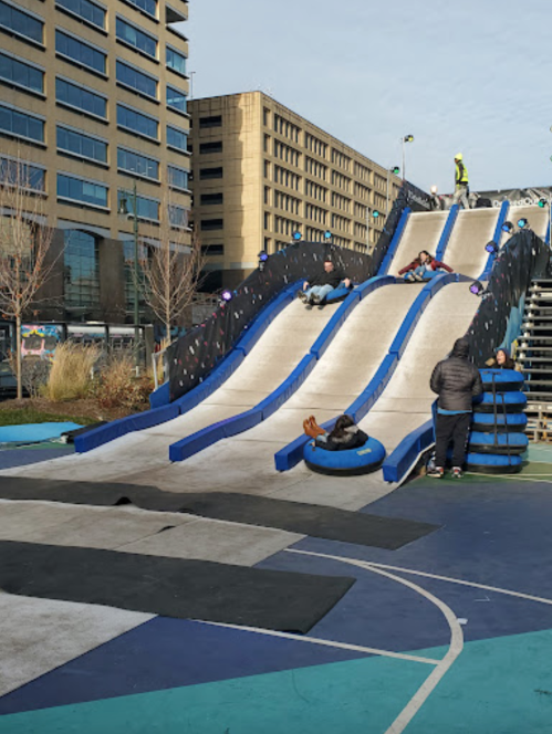 A large, blue and white slide with people sliding down, surrounded by a playground area and buildings in the background.