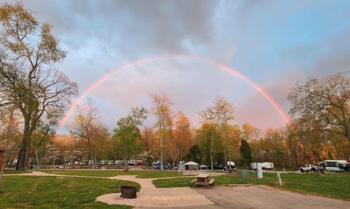 A vibrant rainbow arcs over a campsite surrounded by trees and a cloudy sky, with picnic tables in the foreground.