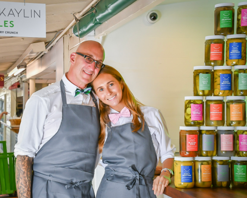 A smiling couple in aprons stands in front of a colorful display of jars filled with pickles.