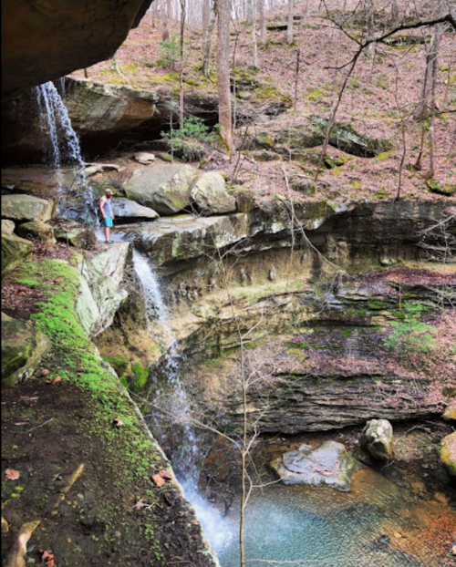 A person stands near a waterfall in a forested area, surrounded by rocks and greenery.