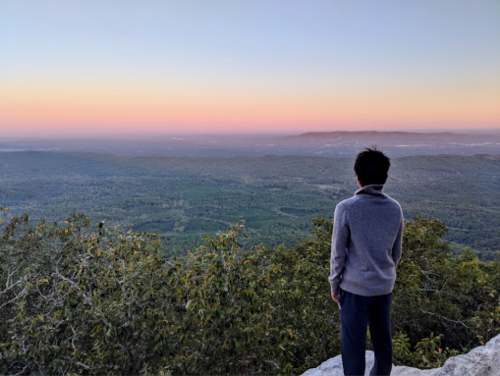 A person stands on a rocky ledge, gazing at a colorful sunset over a vast green landscape.
