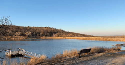 A serene lake scene with a dock, bench, and a hillside under a clear blue sky.