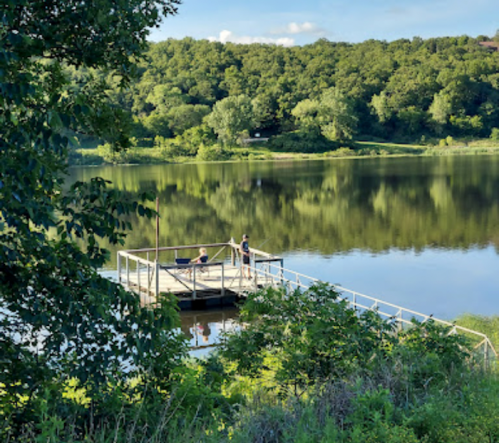 Two people stand on a wooden dock by a calm lake, surrounded by lush greenery and reflecting trees.