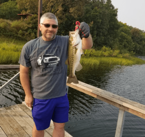 A man in a gray t-shirt and blue shorts holds a large fish while standing on a wooden dock by a lake.