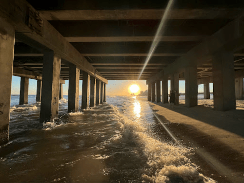 Sunset view under a pier, with sunlight streaming through columns and waves gently lapping at the shore.