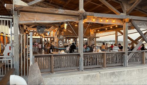 A lively outdoor restaurant with string lights, wooden decor, and diners enjoying their meals at sunset.