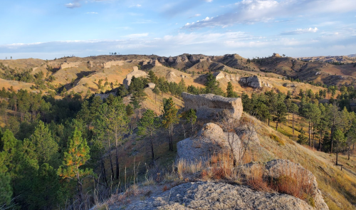 A scenic view of rolling hills and rocky formations, dotted with pine trees under a blue sky with scattered clouds.