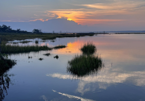 A serene sunset over a calm waterway, with reflections and grassy patches in the foreground.