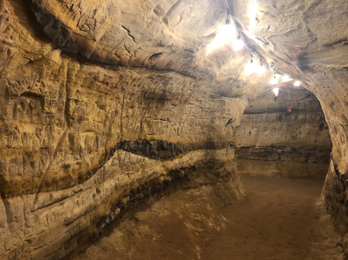 A dimly lit cave interior with carved walls and sandy floor, illuminated by overhead lights.