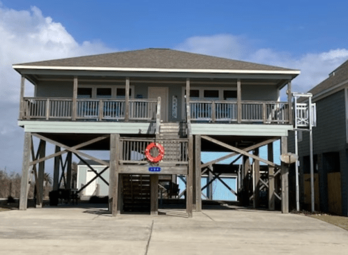 A two-story beach house on stilts with a front porch, surrounded by a concrete driveway and blue sky.