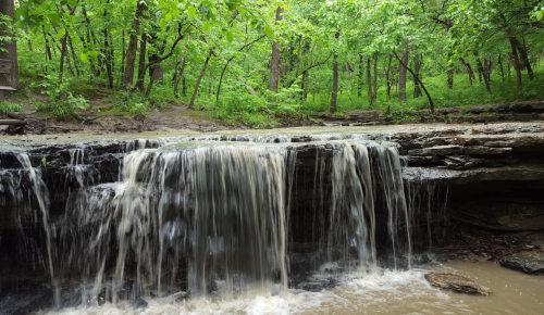 A serene waterfall cascades over rocky ledges, surrounded by lush green trees in a tranquil forest setting.