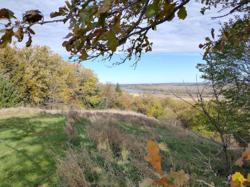 A scenic view of a river valley surrounded by trees and autumn foliage under a partly cloudy sky.