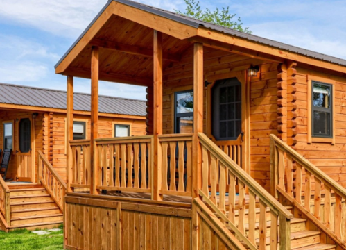 A wooden cabin with a porch, featuring a sloped roof and surrounded by greenery.