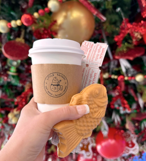 A hand holds a coffee cup and a fish-shaped pastry in front of a festive, decorated background.