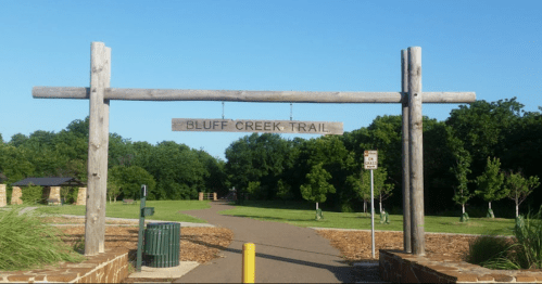 Sign for Bluff Creek Trail at the entrance, surrounded by greenery and a clear blue sky. Pathway visible ahead.