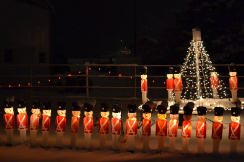 A row of illuminated toy soldiers in red uniforms stands in the snow, with a lit Christmas tree in the background.
