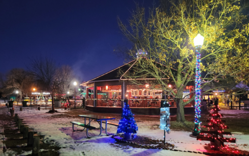 A festive park at night, featuring colorful lights on trees and a cozy building, with snow on the ground.