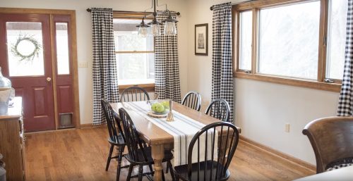 A cozy dining room with a wooden table, black chairs, and checkered curtains, featuring a door and large windows.