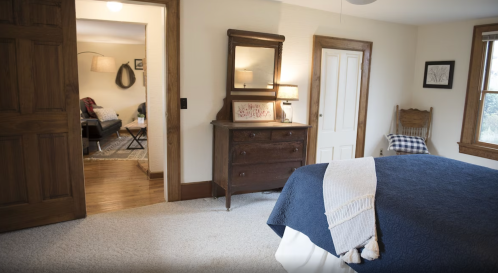 Cozy bedroom with a blue bedspread, wooden furniture, and a doorway leading to a living area. Natural light filters in.