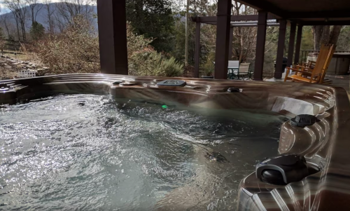 A hot tub with bubbling water, surrounded by trees and a patio area with chairs in the background.
