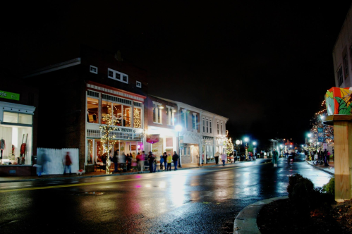 A quiet, illuminated street at night, lined with shops and festive lights reflecting on wet pavement.