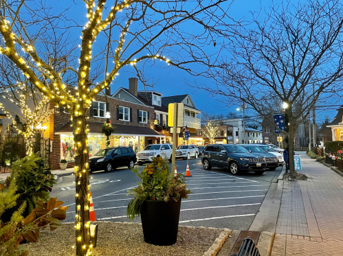 A charming street scene at dusk, featuring lit trees, parked cars, and quaint shops along a cozy sidewalk.