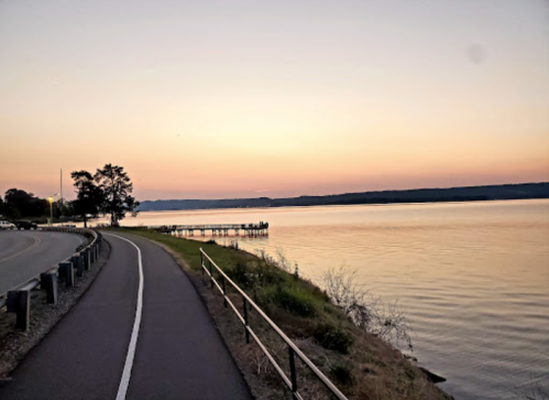 A serene lakeside view at sunset, with a path, trees, and a dock extending into the calm water.