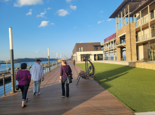 Two people walk along a wooden boardwalk by the water, with a modern building and green lawn in the background.