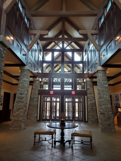 Spacious lobby with high wooden ceilings, stone pillars, and large windows, featuring a central table and chairs.