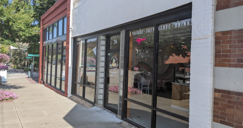 Exterior view of a storefront with large glass windows, featuring signage for soft serve drinks and a flower-lined sidewalk.