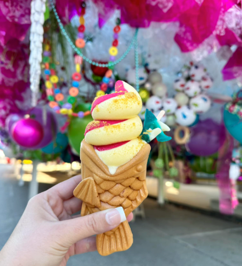 A hand holds a colorful ice cream cone shaped like a fish, with vibrant decorations in the background.