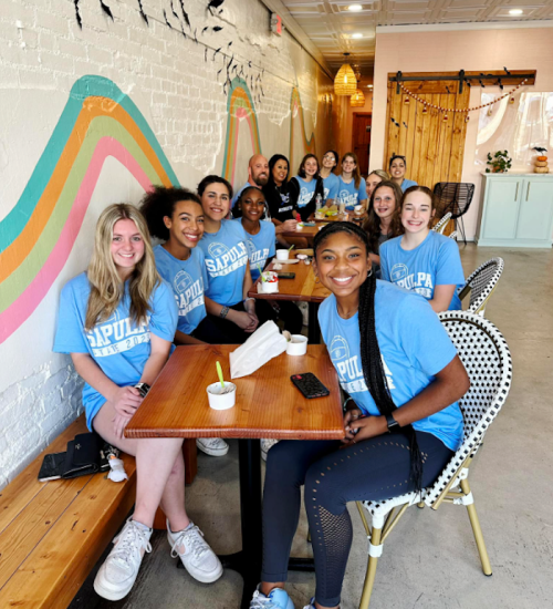 A group of diverse young people in matching blue shirts sitting at a table in a bright, casual café.