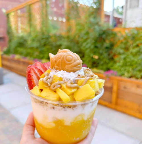 A colorful dessert cup with mango, coconut, and a fish-shaped cookie on top, held in a hand against a green background.